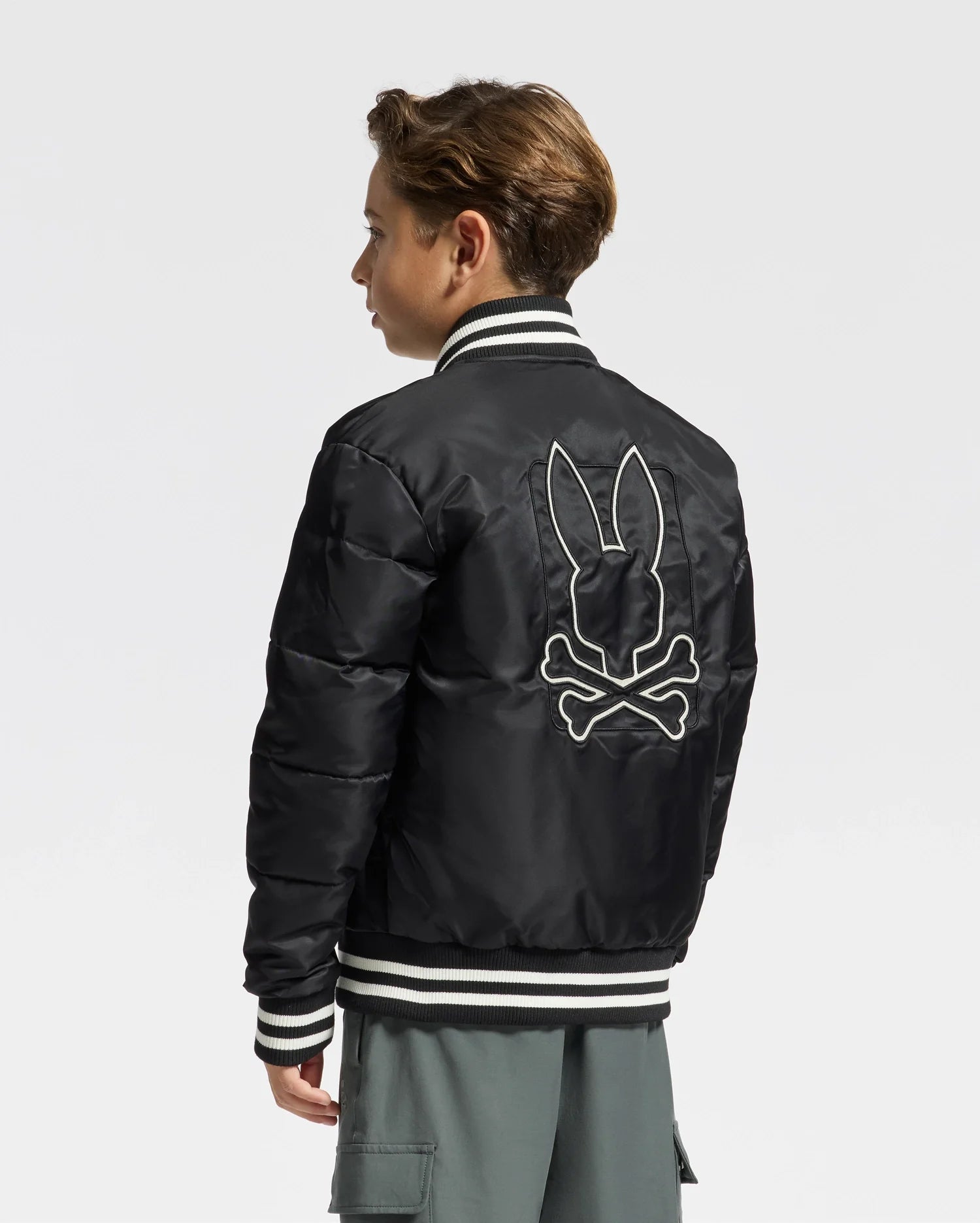A person is sporting the stylish KIDS FRASER FASHION PUFFER by Psycho Bunny, a black puffer jacket featuring white striped cuffs and a graphic bunny skull and crossbones on the back. They are posed sideways, gazing to their left against a simple white background.