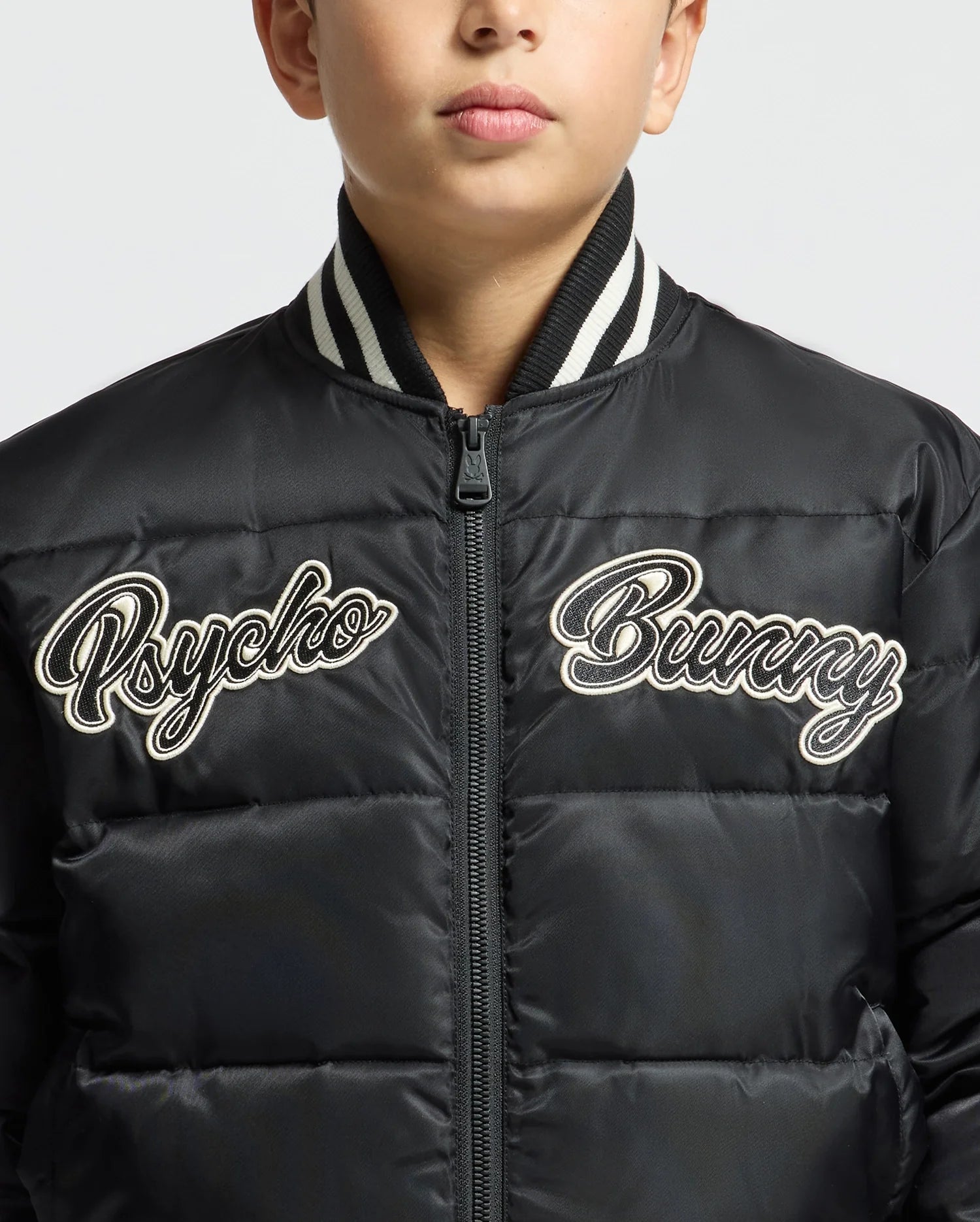 A boy wears the KIDS FRASER FASHION PUFFER - B0N338E200 from Psycho Bunny, a trendy black puffer jacket with white embroidered text "Psycho Bunny" on the front. This warm kids' fashion piece, featuring a ribbed collar, keeps him cozy. His face is partially visible against a plain background.