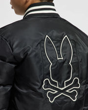 A person is wearing the Psycho Bunny KIDS FRASER FASHION PUFFER in black, which features a trendy large embroidery of a rabbit head silhouette with crossbones underneath. The jacket is reminiscent of stylish kids' puffers and includes a ribbed collar with black and white stripes, infusing playful sophistication.