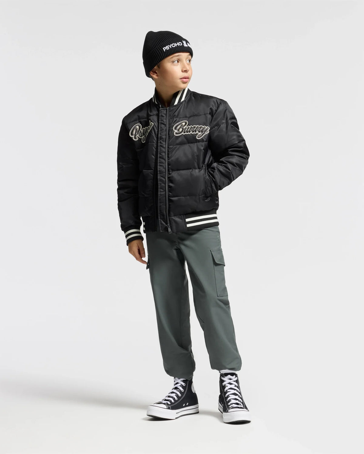 A young boy strikes a pose against a plain backdrop, wearing the KIDS FRASER FASHION PUFFER - B0N338E200 by Psycho Bunny, featuring embroidered text. Completing his outfit are a black beanie, green cargo pants, and stylish high-top sneakers. He looks to the side with one hand casually in his pocket.