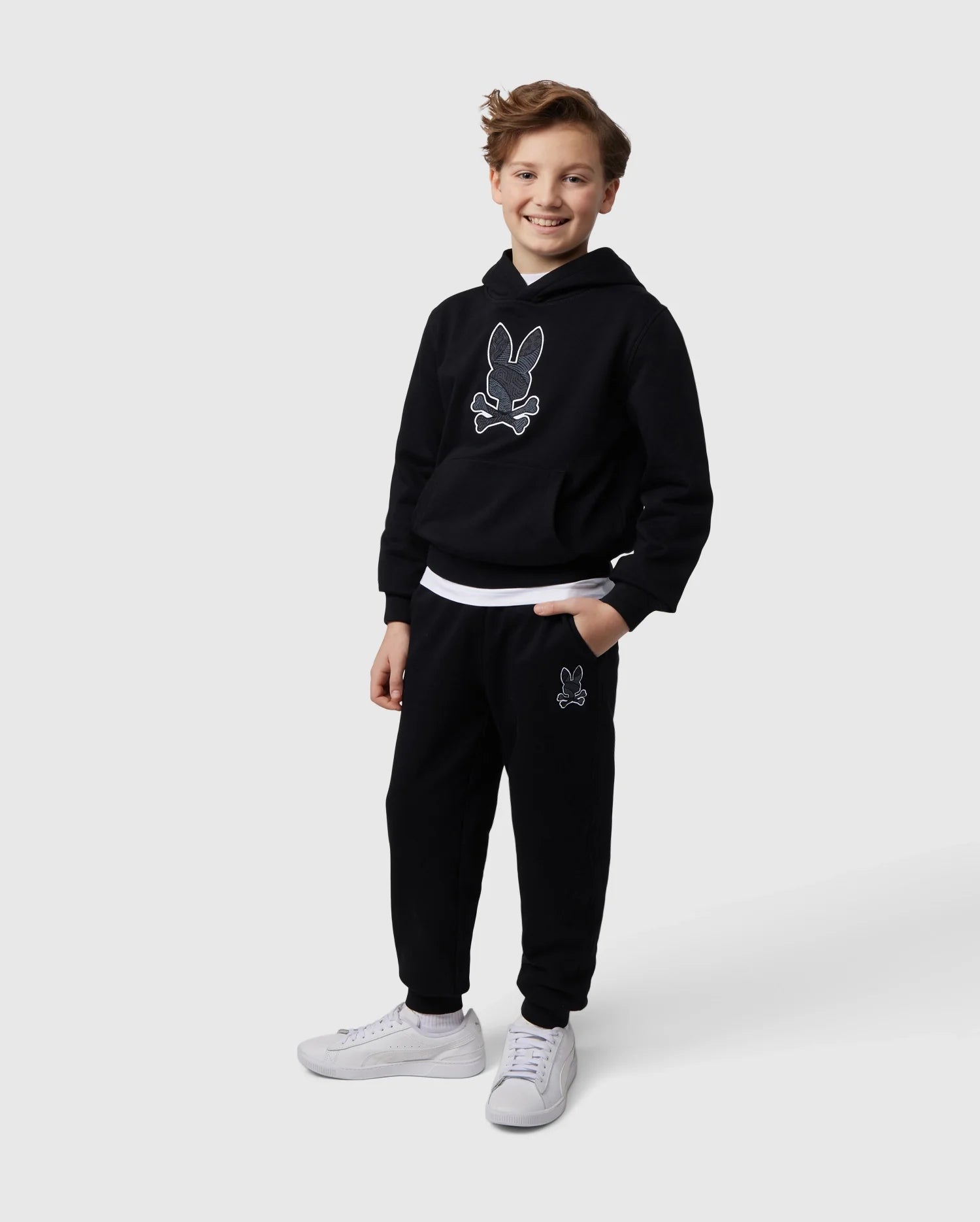 A smiling young person stands against a plain white background, wearing the KIDS LENOX REGULAR FIT SWEATPANT and a matching black hoodie featuring an embroidered bunny logo from Psycho Bunny. They have short, wavy hair and wear white sneakers. One hand is in their pocket, and they appear relaxed and happy.