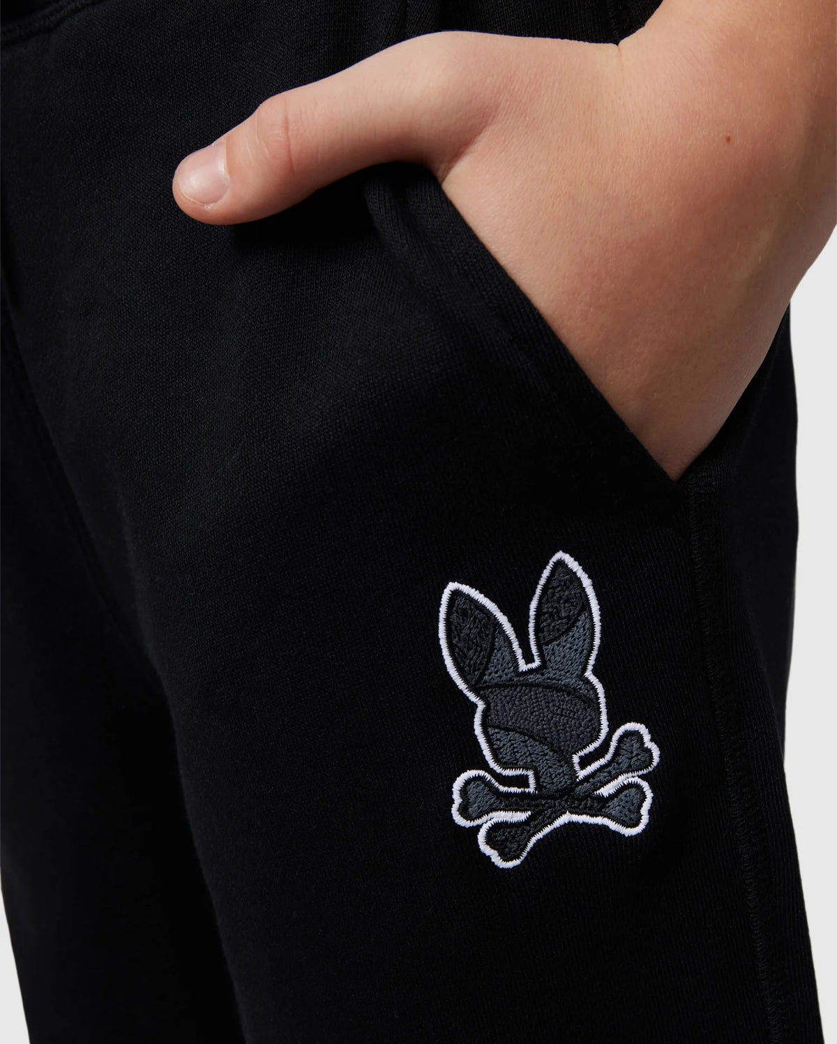A close-up image of a person with their right hand placed inside the pocket of Psycho Bunny's KIDS LENOX REGULAR FIT SWEATPANT in black. The pants feature an embroidered logo near the pocket depicting a black and white skull and crossbones with bunny ears.