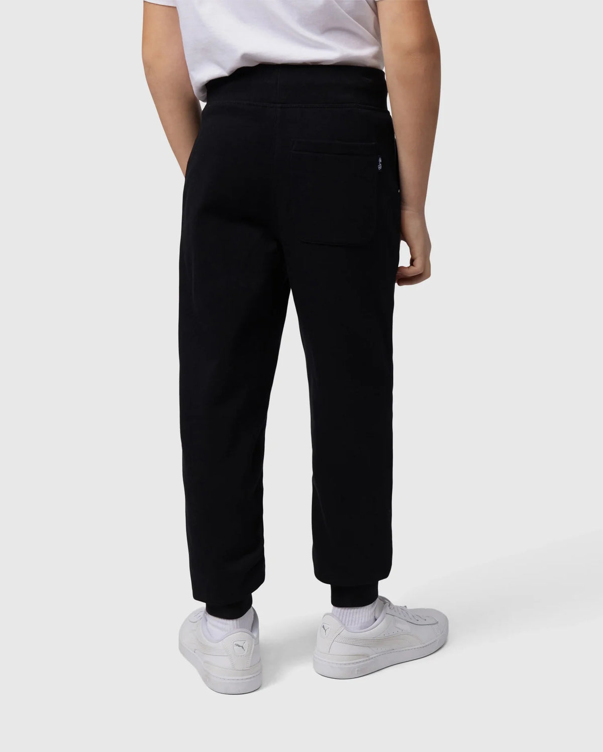 A back view of a person wearing a white T-shirt, white sneakers, and Psycho Bunny's KIDS LENOX REGULAR FIT SWEATPANT in black made from soft cotton fleece with an embroidered logo on the back pocket. The person's right arm is resting by their side against a plain white background.