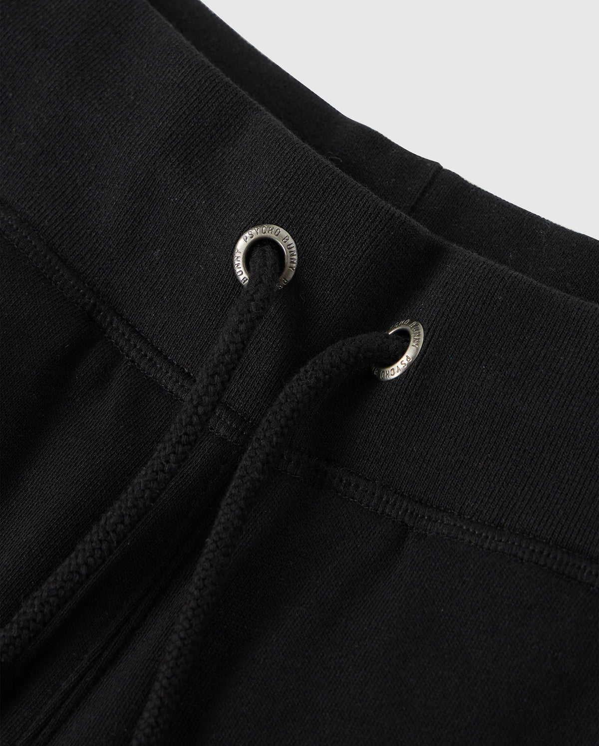 Close-up of the waistband of the Psycho Bunny KIDS LENOX REGULAR FIT SWEATPANT in black. The drawstring is threaded through metal eyelets, with the ends of the strings visible. Made from soft cotton fleece, typical of casual or athletic wear. The background is a plain light grey.