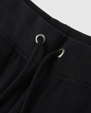 Close-up of the waistband of the Psycho Bunny KIDS LENOX REGULAR FIT SWEATPANT in black. The drawstring is threaded through metal eyelets, with the ends of the strings visible. Made from soft cotton fleece, typical of casual or athletic wear. The background is a plain light grey.