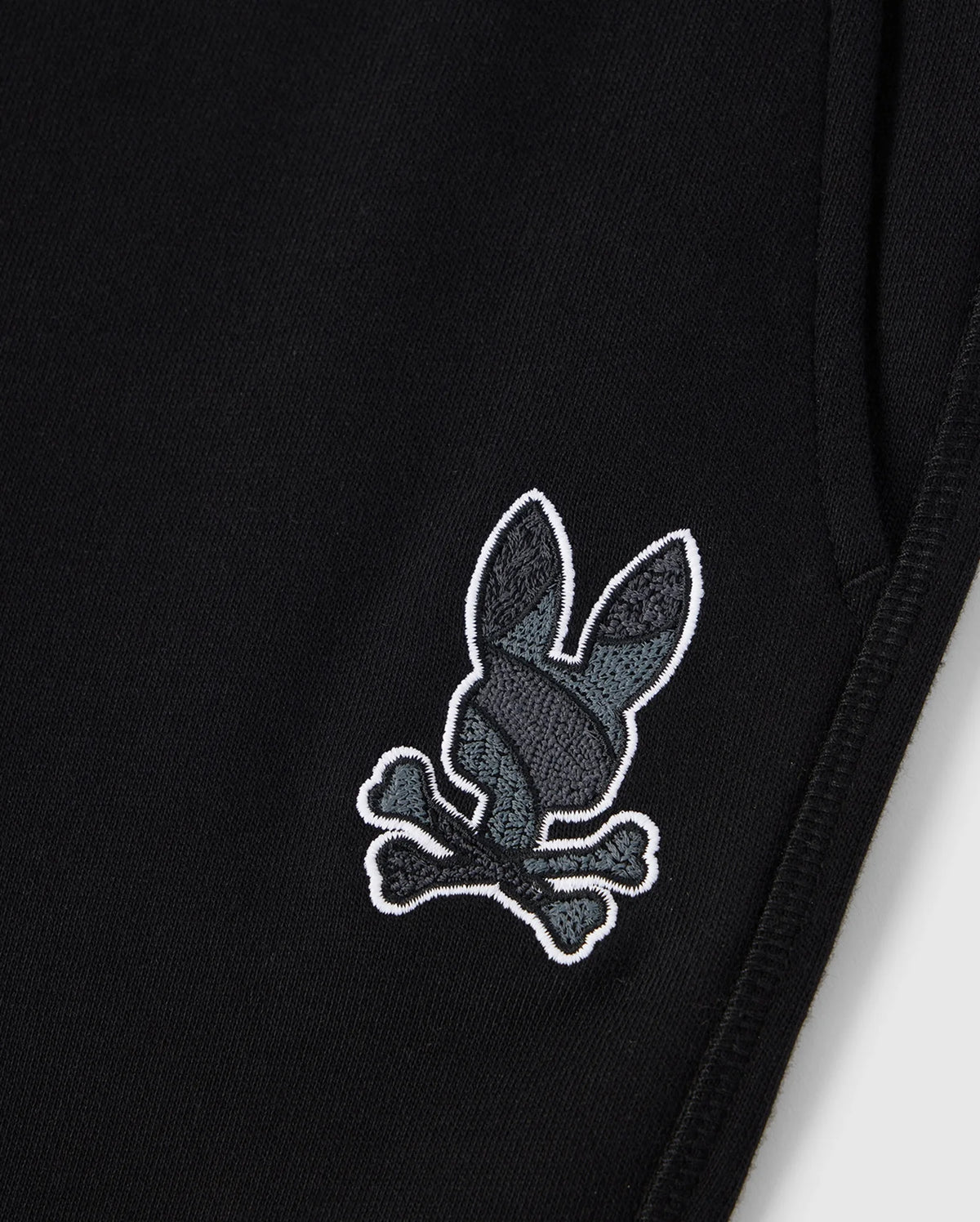 A close-up view of the KIDS LENOX REGULAR FIT SWEATPANT by Psycho Bunny, in black cotton fleece. The sweatpants feature an embroidered logo of a gray rabbit head with black detailing and crossed bones underneath, mimicking the traditional pirate skull and crossbones symbol.