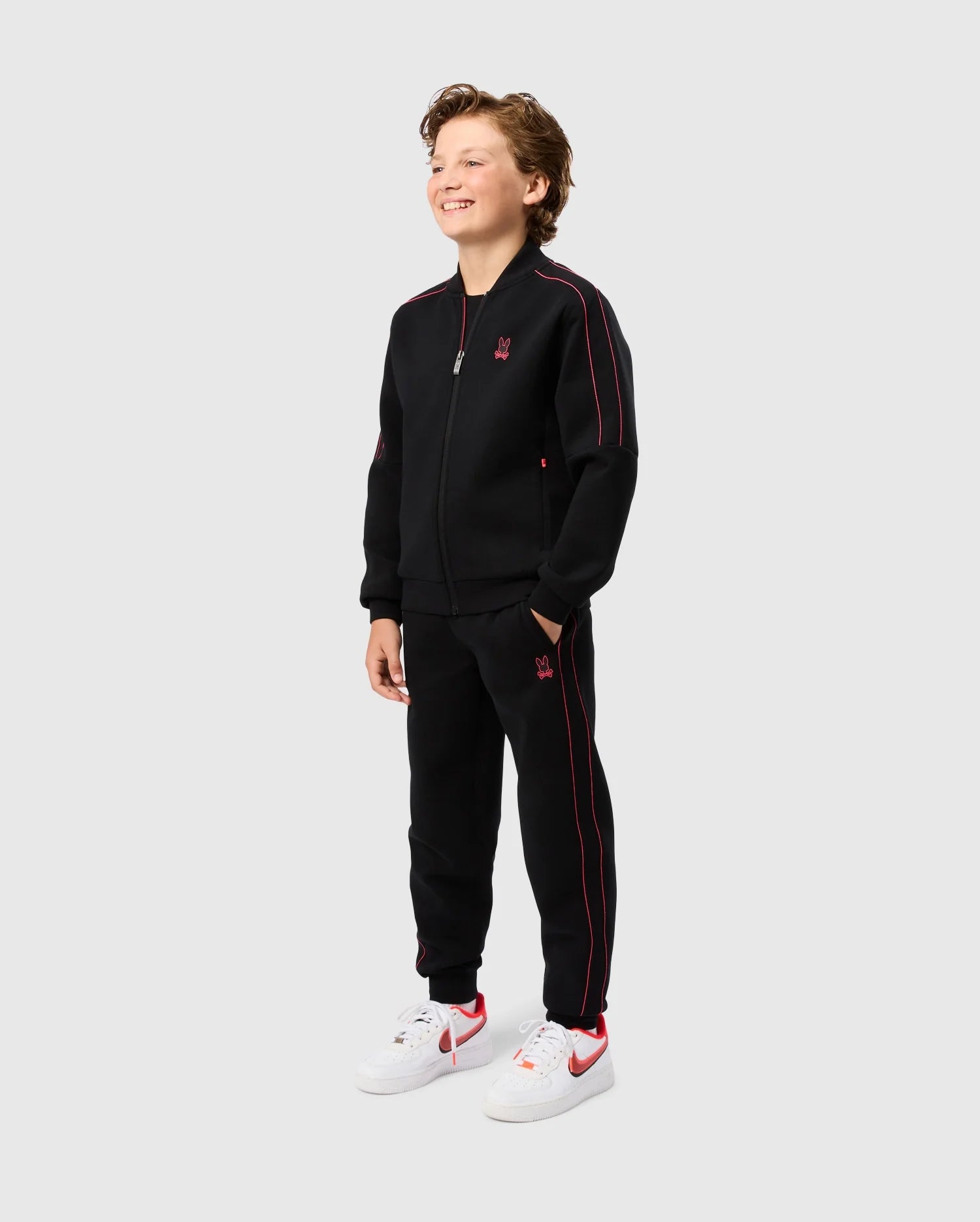 A young boy stands smiling, dressed in the KIDS TRAVIS TRACK PANT - B0P143D200 from Psycho Bunny. He has his hands in the elastic and comfortable pants pockets of his black zip-up tracksuit with red accents. The outfit features a small red logo on the chest and thigh. He complements his look with white sneakers featuring red details, all against a plain grey background.