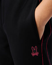 Close-up of a person wearing the KIDS TRAVIS TRACK PANT (B0P143D200) by Psycho Bunny, with their hand in the pocket. The black pants feature a red bunny and crossed bones logo near the pocket, along with red stitching along the seam. These elastic and comfortable kids' track pants also sport contrast chainstitch stripes for an extra touch of style.