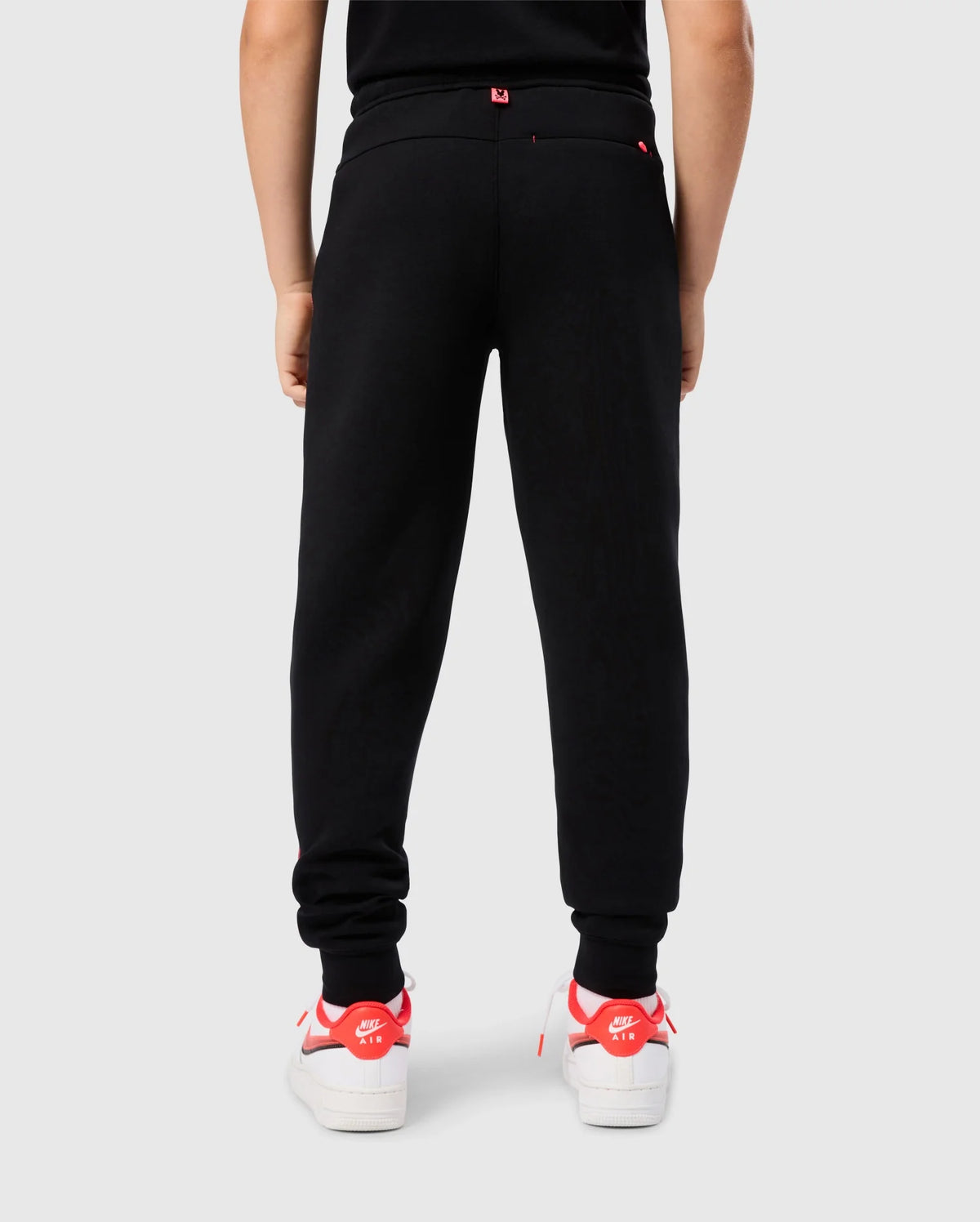 A person is standing with their back to the camera, wearing the black, elastic and comfortable KIDS TRAVIS TRACK PANT - B0P143D200 by Psycho Bunny and white sneakers with red accents. The individual appears to be against a plain white background.