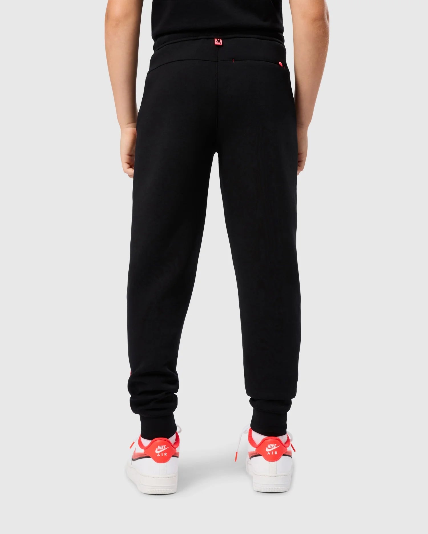 A person is standing with their back to the camera, wearing the black, elastic and comfortable KIDS TRAVIS TRACK PANT - B0P143D200 by Psycho Bunny and white sneakers with red accents. The individual appears to be against a plain white background.