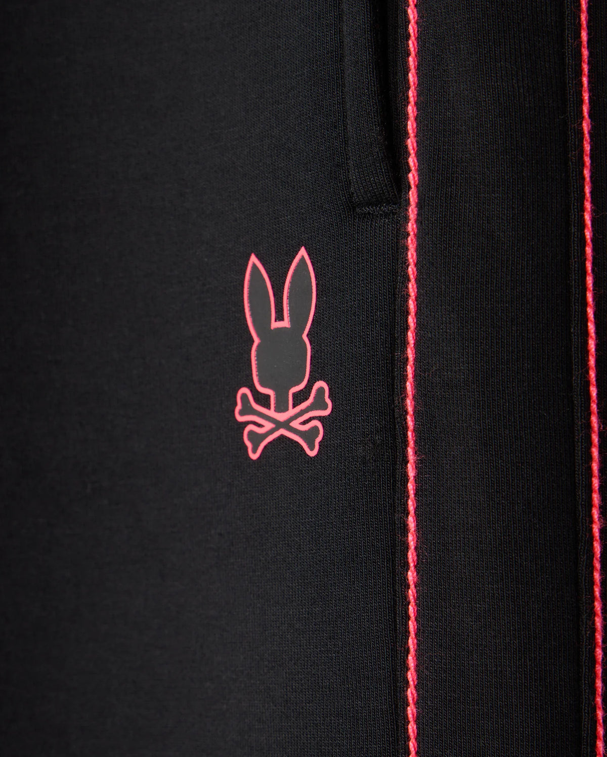 Close-up of the black fabric of the KIDS TRAVIS TRACK PANT - B0P143D200 by Psycho Bunny, featuring a pink bunny skull and crossbones logo. The elastic and comfortable fabric showcases contrast chainstitch stripes running vertically to the right of the logo.