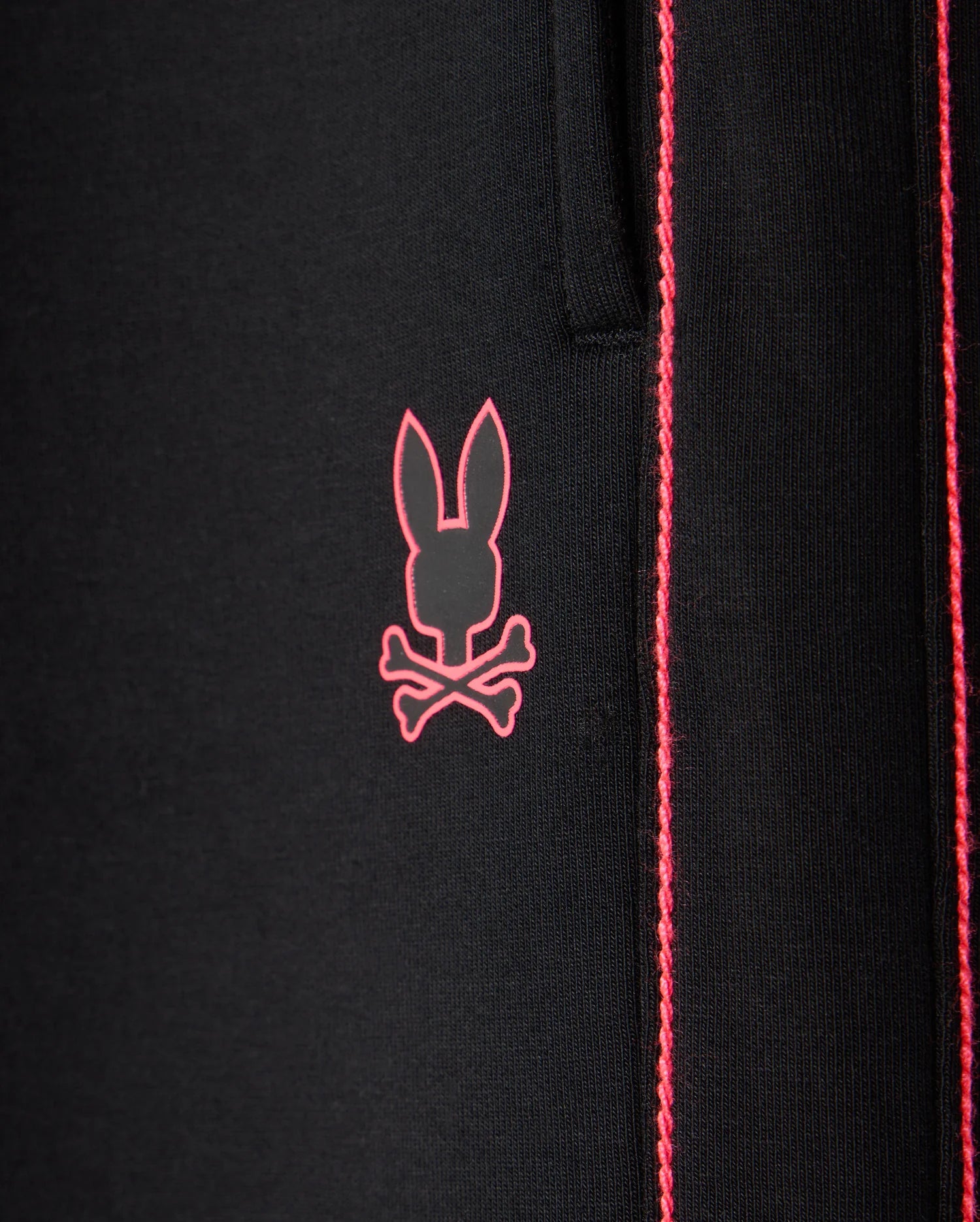 Close-up of the black fabric of the KIDS TRAVIS TRACK PANT - B0P143D200 by Psycho Bunny, featuring a pink bunny skull and crossbones logo. The elastic and comfortable fabric showcases contrast chainstitch stripes running vertically to the right of the logo.