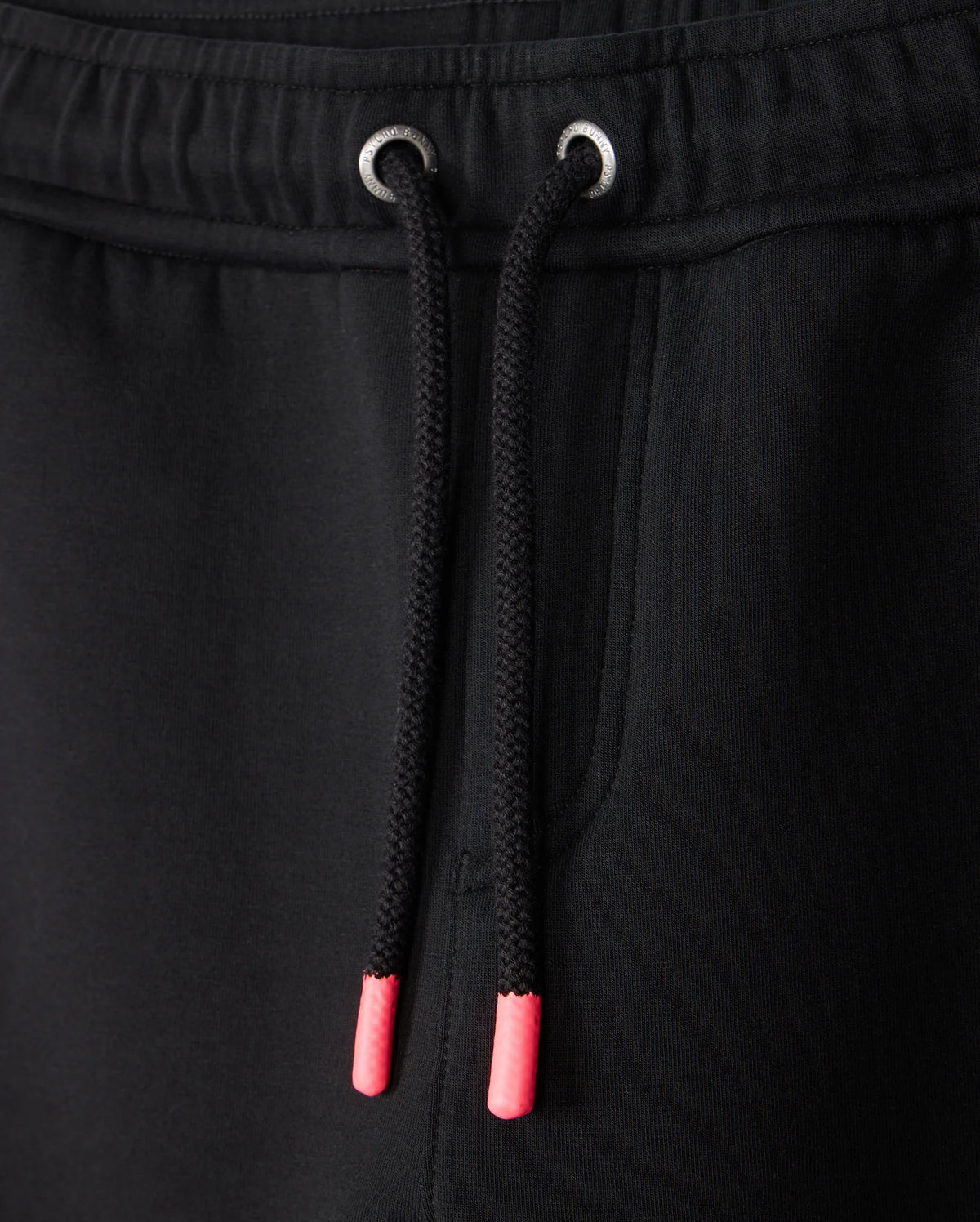 Close-up of the KIDS TRAVIS TRACK PANT - B0P143D200 by Psycho Bunny, showcasing black jogger pants with a drawstring featuring pink plastic aglets and metal eyelets, complemented by a side pocket. The elastic and comfortable material makes it ideal for kids' track pants.