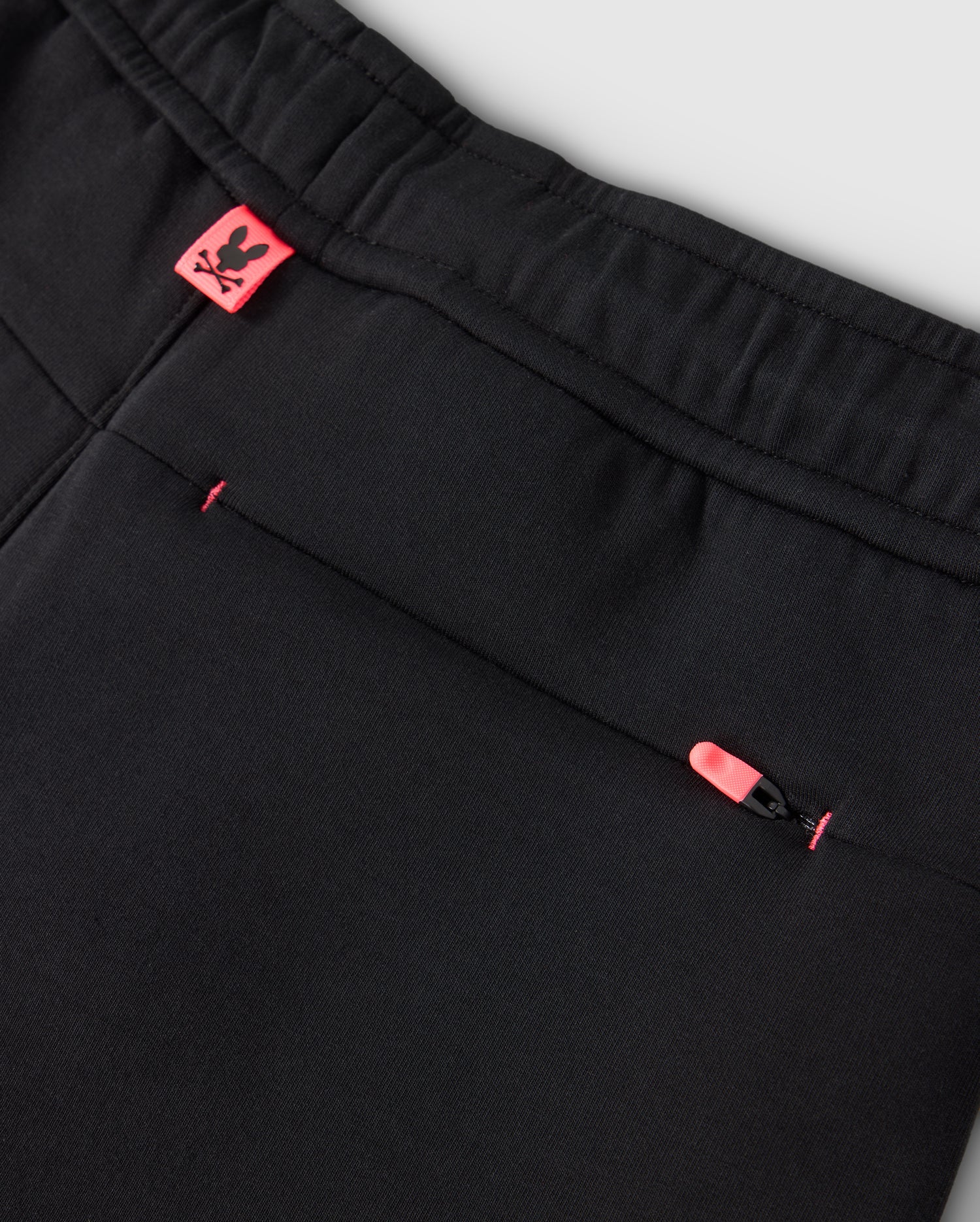Close-up of the KIDS TRAVIS TRACK PANT - B0P143D200 by Psycho Bunny, showcasing an elastic and comfortable waistband. These black pants include a back pocket with a pink zipper pull and a small pink tag with the Psycho Bunny logo near the waistband. The fabric is smooth and well-stitched, accented by contrast chainstitch stripes for added style.