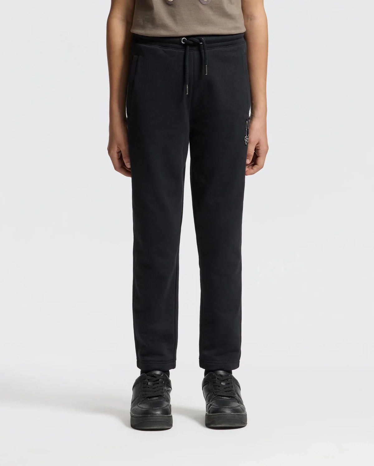 A person wearing the KIDS ALEXANDER SWEATPANT - B0P155E200 by Psycho Bunny in black, paired with black sneakers, stands against a plain white background. The comfortable jogging pants enhance the look of the partially visible upper body, which is adorned with a brown top.