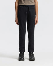 A person wearing the KIDS ALEXANDER SWEATPANT - B0P155E200 by Psycho Bunny in black, paired with black sneakers, stands against a plain white background. The comfortable jogging pants enhance the look of the partially visible upper body, which is adorned with a brown top.