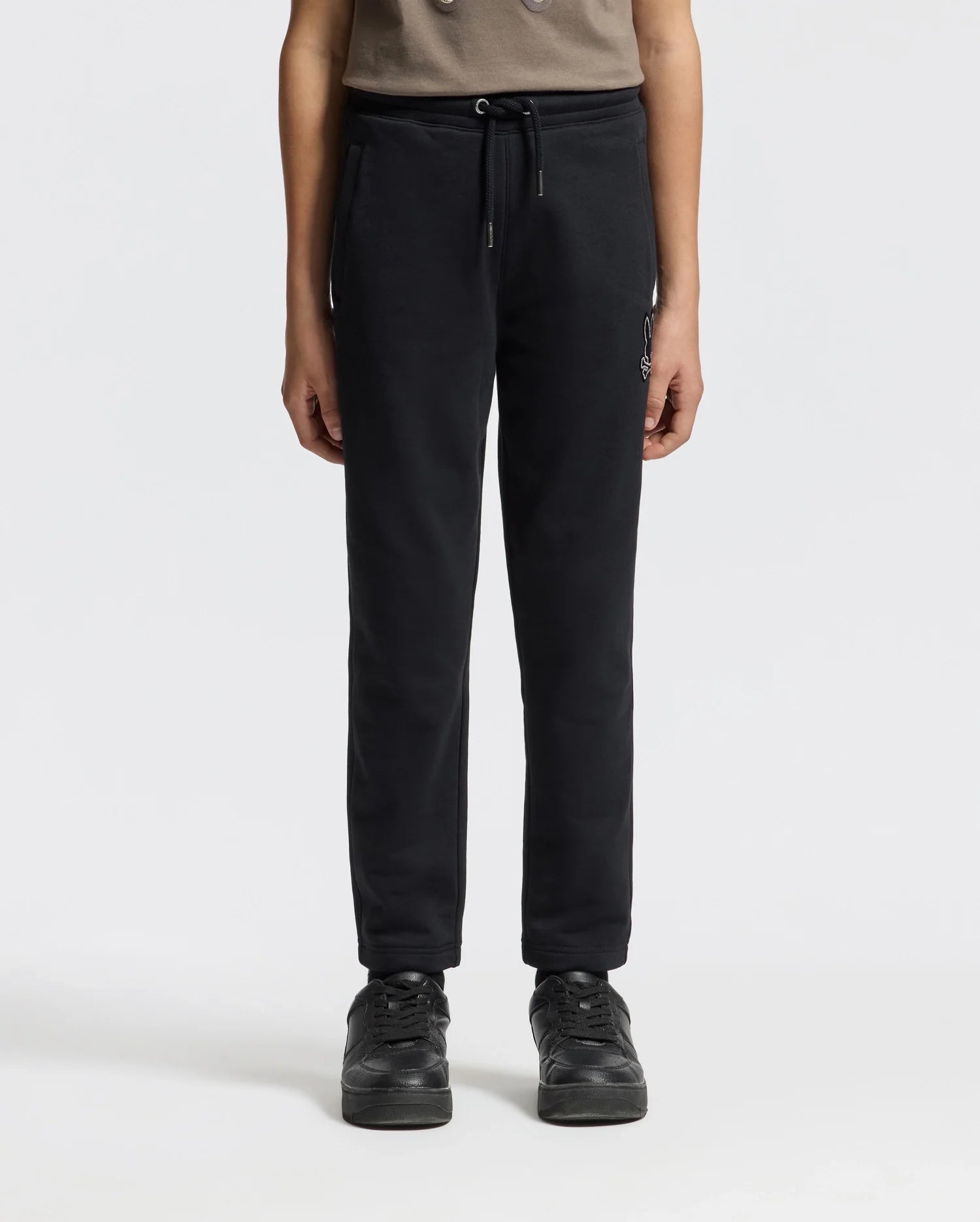 A person wearing the KIDS ALEXANDER SWEATPANT - B0P155E200 by Psycho Bunny in black, paired with black sneakers, stands against a plain white background. The comfortable jogging pants enhance the look of the partially visible upper body, which is adorned with a brown top.
