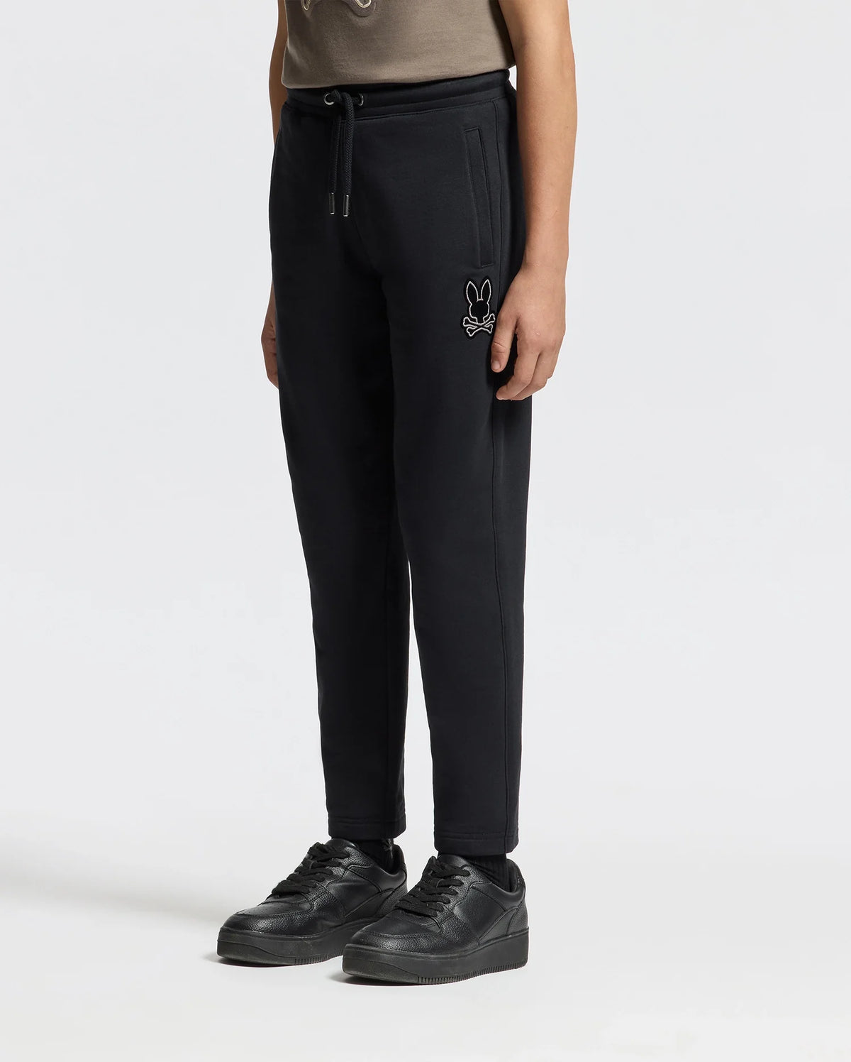 A child wearing the KIDS ALEXANDER SWEATPANT - B0P155E200 by Psycho Bunny, featuring a small embroidered cartoon logo, pairs them with black sneakers while standing against a plain white background. Only the lower half of their body is visible.