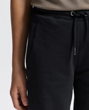A person is wearing the Psycho Bunny KIDS ALEXANDER SWEATPANT - B0P155E200 in black, featuring a side pocket. Their right hand is partially visible, resting by their side. The sweatpants are paired with a beige top, all set against a plain white background.