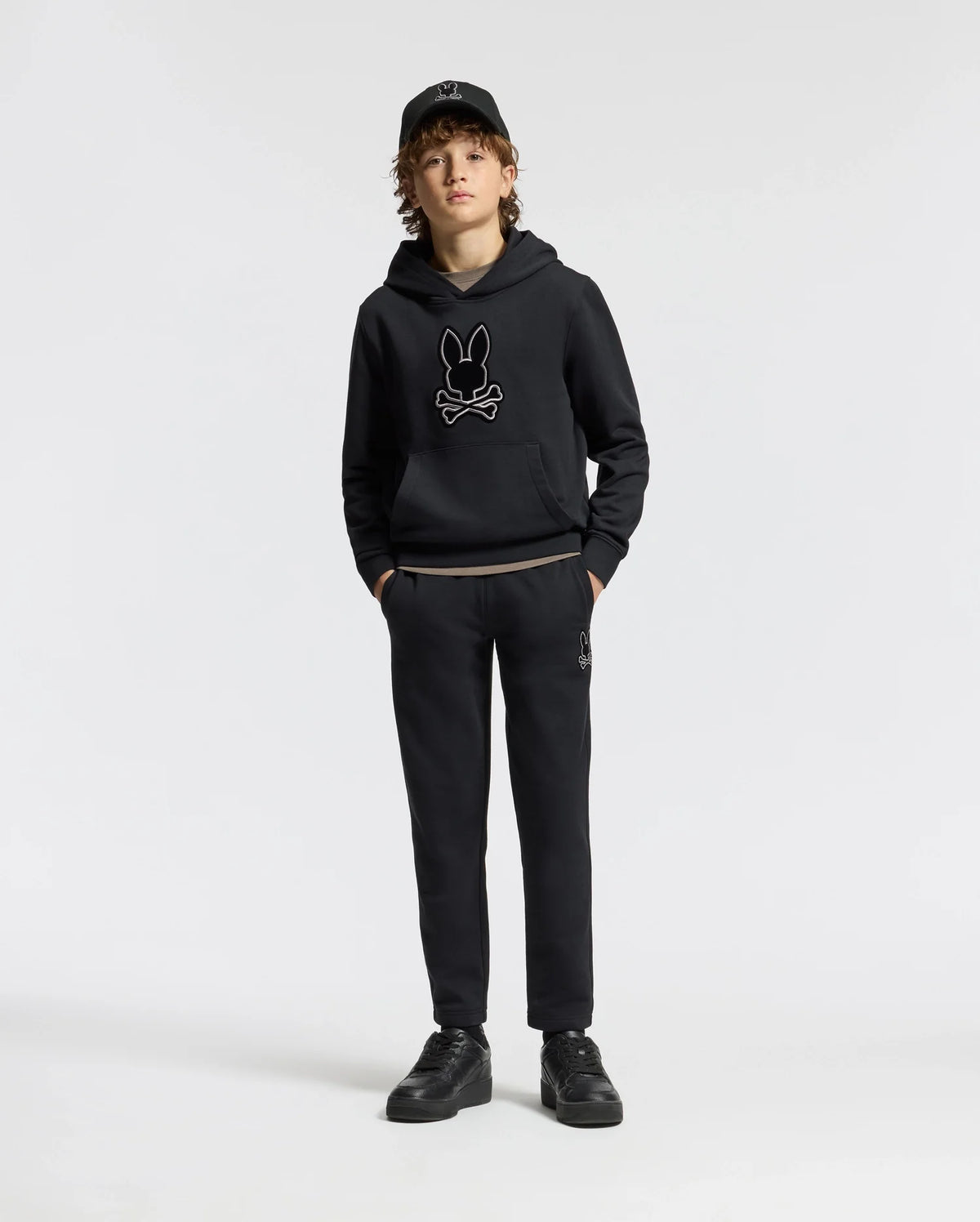 A young person stands confidently, dressed in a black hoodie featuring a rabbit skull design, kids' black sweatpants from the Psycho Bunny collection, a matching cap, and sneakers. The sleek cotton blend of the KIDS ALEXANDER SWEATPANT - B0P155E200 enhances their edgy style against the plain white background.