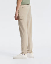 A person wearing Psycho Bunny's KIDS GREEN VALLEY CARGO PANT - B0P199E200 in beige and white sneakers stands against a plain background. These pants offer a stylish yet functional design with their comfortable fit and ample cargo pockets. They wear a light green top, partially visible.