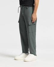 The image depicts a person from the waist down wearing the KIDS GREEN VALLEY CARGO PANT - B0P199E200 by Psycho Bunny in dark green, paired with white sneakers. These stylish kids' trousers feature large pockets and a drawstring waist, set against a plain white background.