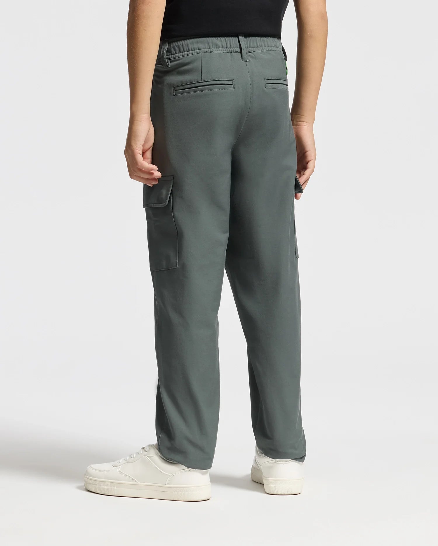 A person wearing the KIDS GREEN VALLEY CARGO PANT - B0P199E200 in a vibrant green stands with their back to the camera, paired with crisp white sneakers. These stylish cargo pants from Psycho Bunny feature side pockets and an elastic waistband. The ensemble is completed with a black top.
