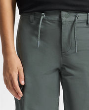 A person is wearing the Psycho Bunny KIDS GREEN VALLEY CARGO PANT - B0P199E200. These trendy dark green cargo pants feature a drawstring, button closure, and belt loops. The focus is on the waist to mid-thigh area, where a hand rests beside the pants, emphasizing their contemporary style.