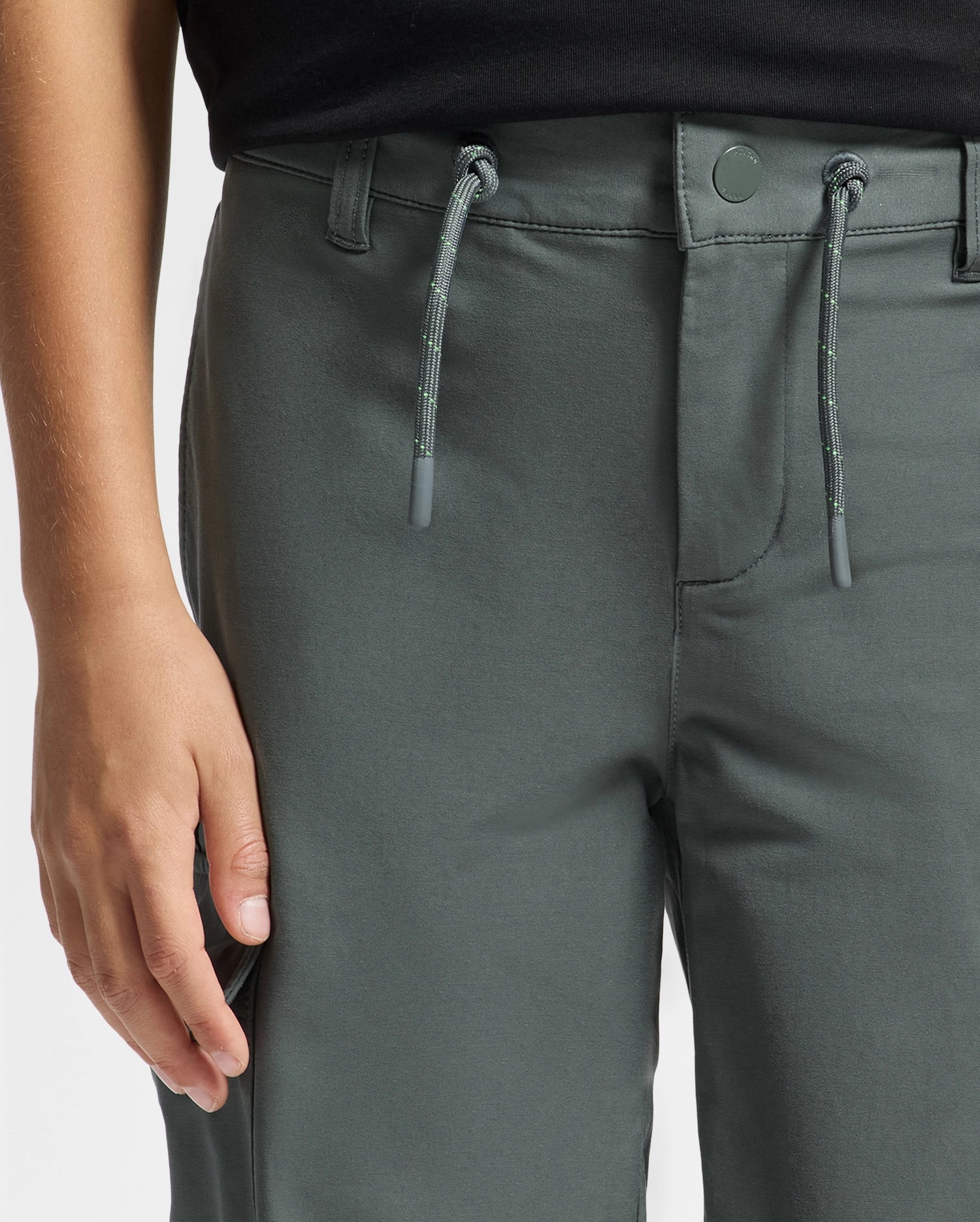 A person is wearing the Psycho Bunny KIDS GREEN VALLEY CARGO PANT - B0P199E200. These trendy dark green cargo pants feature a drawstring, button closure, and belt loops. The focus is on the waist to mid-thigh area, where a hand rests beside the pants, emphasizing their contemporary style.