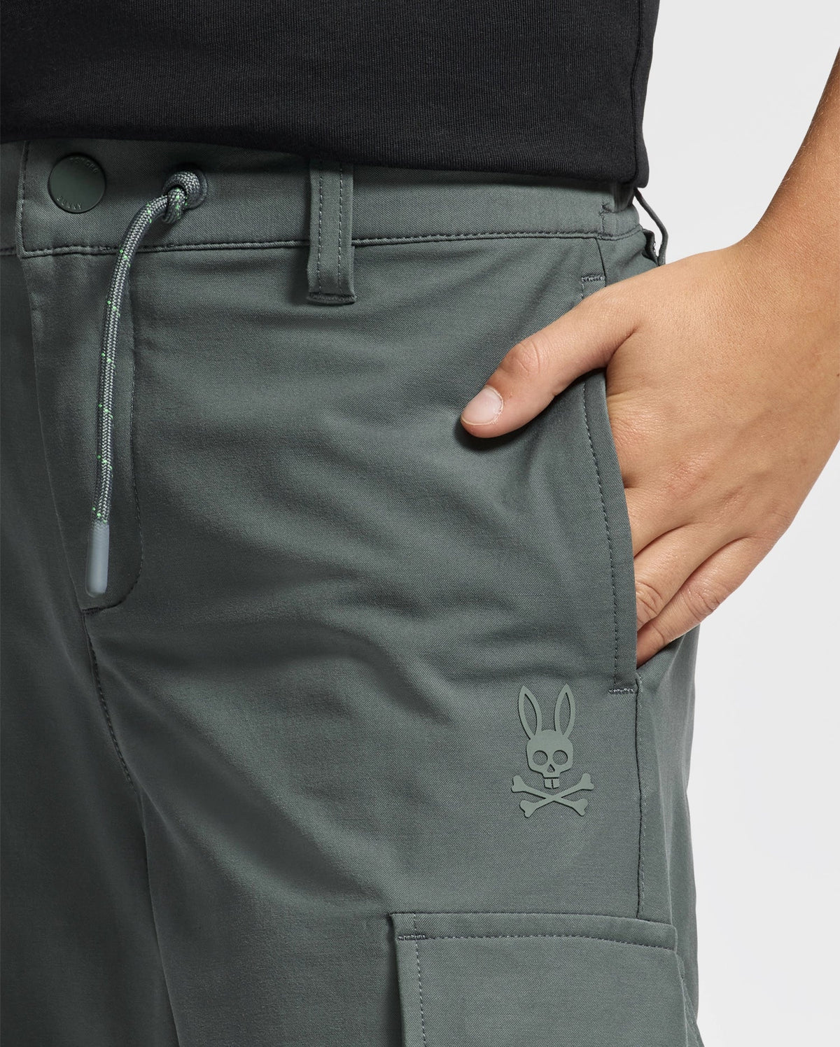 Close-up of a person wearing the fashionable KIDS GREEN VALLEY CARGO PANT by Psycho Bunny, paired with a black shirt. Their hand rests in the pocket, which is adorned with a small logo featuring Psycho Bunny's signature rabbit with crossbones.