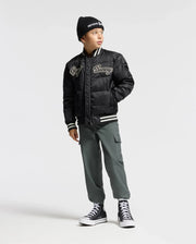 A child wearing a black beanie, a black puffer jacket with white text, and the trendy KIDS GREEN VALLEY CARGO PANT by Psycho Bunny stands against a plain background, looking off to the side in stylish kids' trousers.