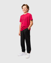 A boy with light brown hair smiles while wearing a bright pink t-shirt and black KIDS MADISON COMMUTER PANT - B0P395Z1WB with zippered expansion vents, his hands in his pockets. He is also sporting white sneakers and a detachable Psycho Bunny keychain. The background is plain and white.