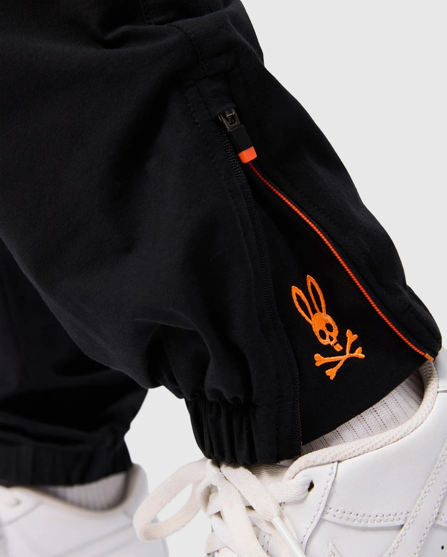Close-up of a child wearing the KIDS MADISON COMMUTER PANT by Psycho Bunny in black, featuring an orange embroidery of a bunny skull and crossbones near the ankle. The pants have zippered expansion vents and come with a detachable Bunny keychain. The child is also sporting white sneakers.