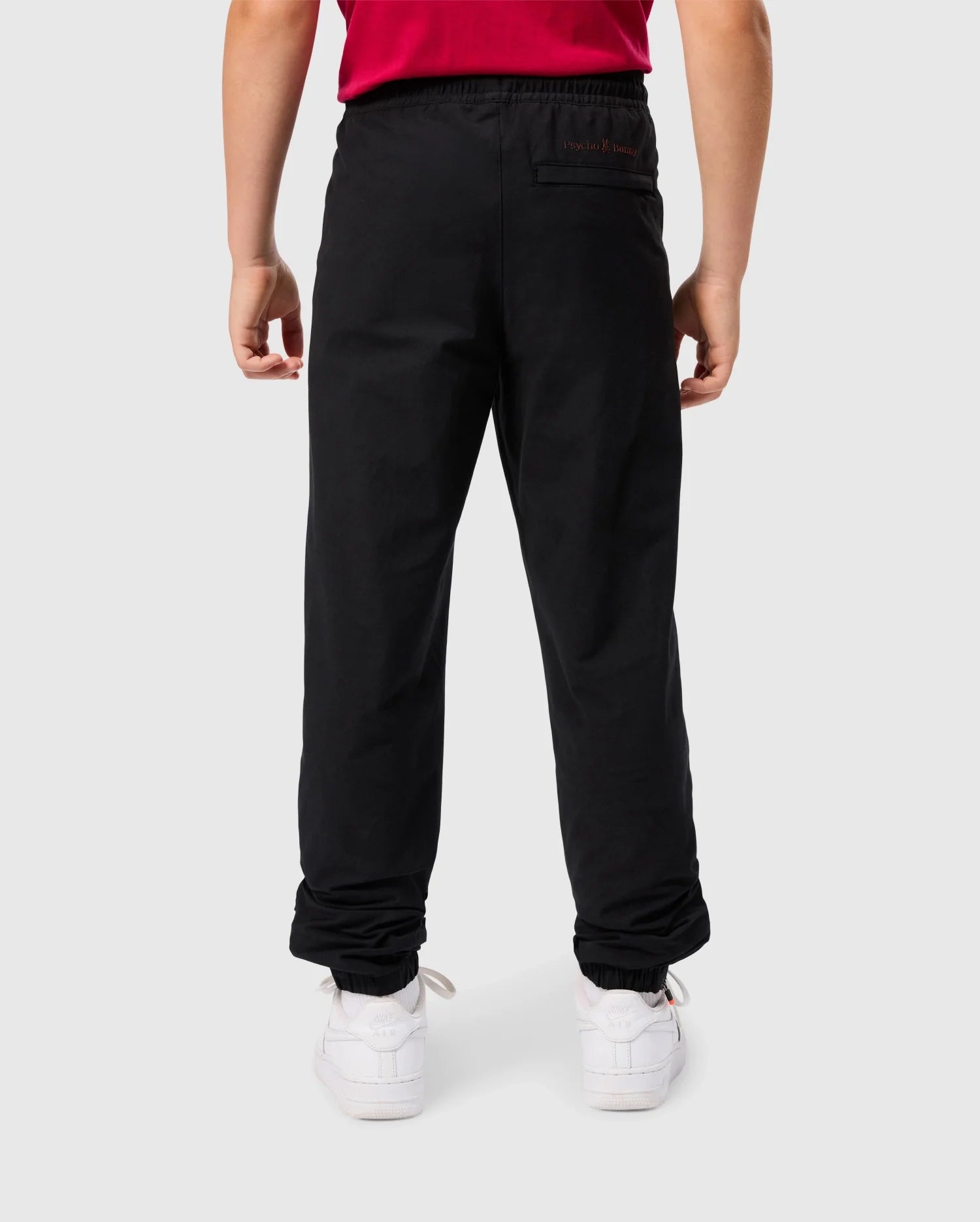 Rear view of a person wearing a red shirt tucked into black KIDS MADISON COMMUTER PANTS by Psycho Bunny, featuring elasticated cuffs and paired with white sneakers. The person stands against a plain light gray background.
