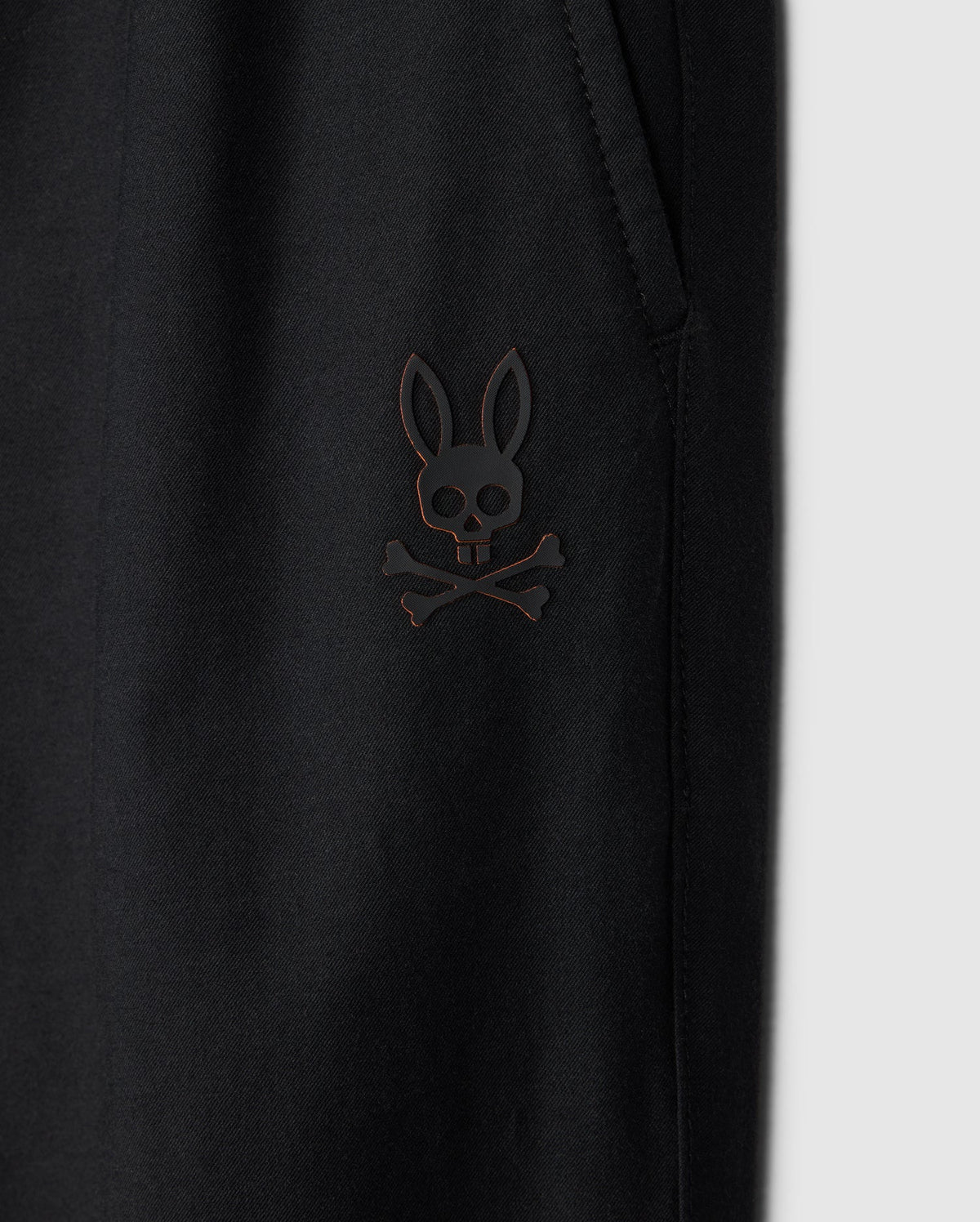 Close-up of a black Psycho Bunny Kids Madison Commuter Pant (B0P395Z1WB) featuring an emblem of a bunny with crossed bones underneath, reminiscent of a skull and crossbones design. The emblem is positioned near a pocket with zippered expansion vents. The background is plain grey.