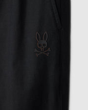 Close-up of a black Psycho Bunny Kids Madison Commuter Pant (B0P395Z1WB) featuring an emblem of a bunny with crossed bones underneath, reminiscent of a skull and crossbones design. The emblem is positioned near a pocket with zippered expansion vents. The background is plain grey.