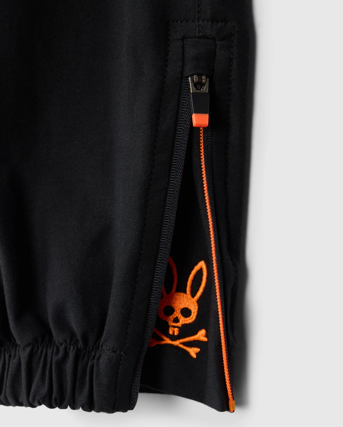 Close-up of the KIDS MADISON COMMUTER PANT by Psycho Bunny in black with an orange zipper. The zipper is partially open, revealing an orange skull and crossbones graphic with bunny ears on the inner fabric. This garment features an elastic cuff at the hem, reminiscent of the Madison commuter pant design.