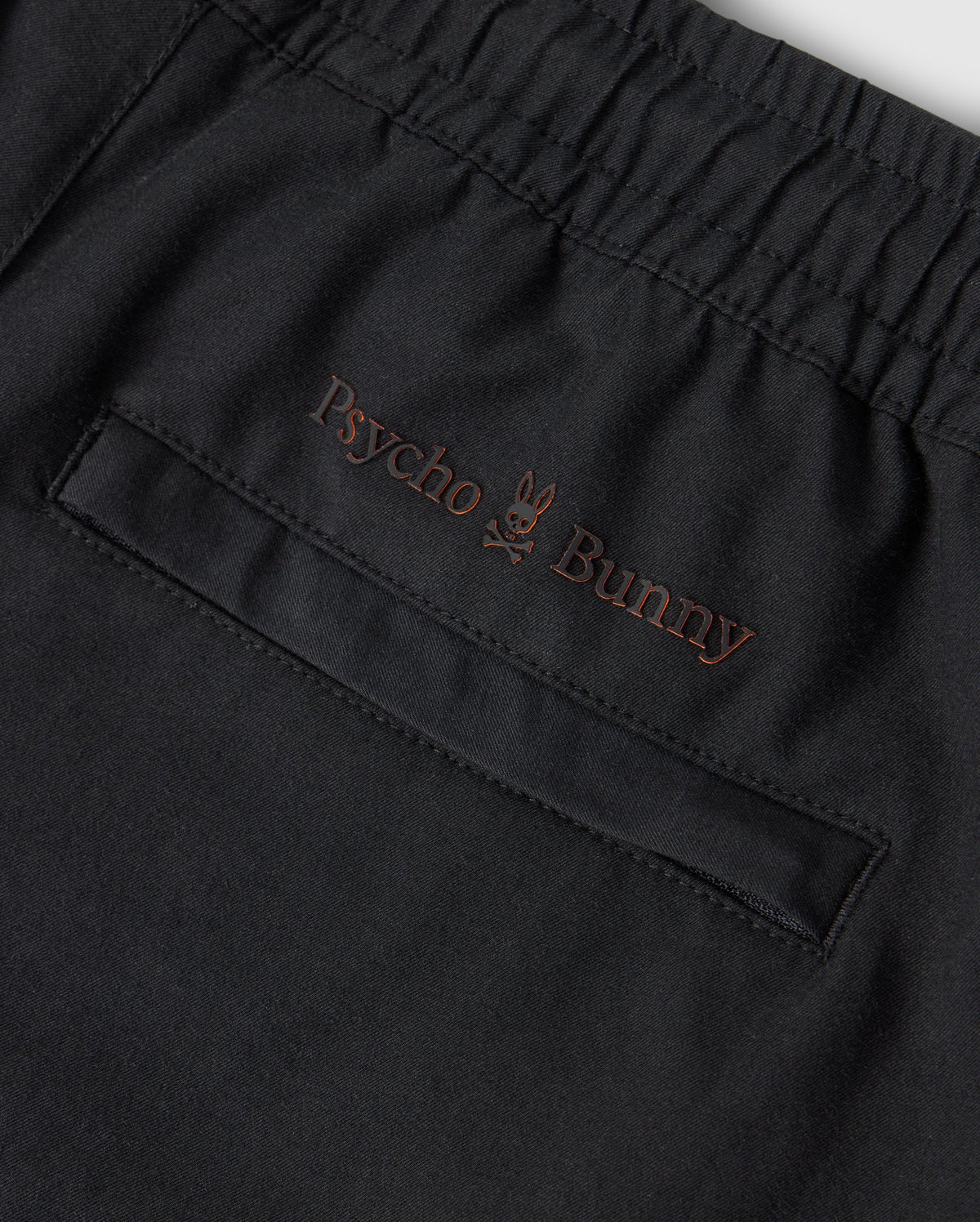 Close-up of a black KIDS MADISON COMMUTER PANT - B0P395Z1WB from Psycho Bunny, featuring an elastic waistband and a pocket with subtle brown embroidery. The embroidery includes the text "Psycho Bunny" with a small bunny logo positioned between the two words. Zippered expansion vents add functionality to this stylish piece.