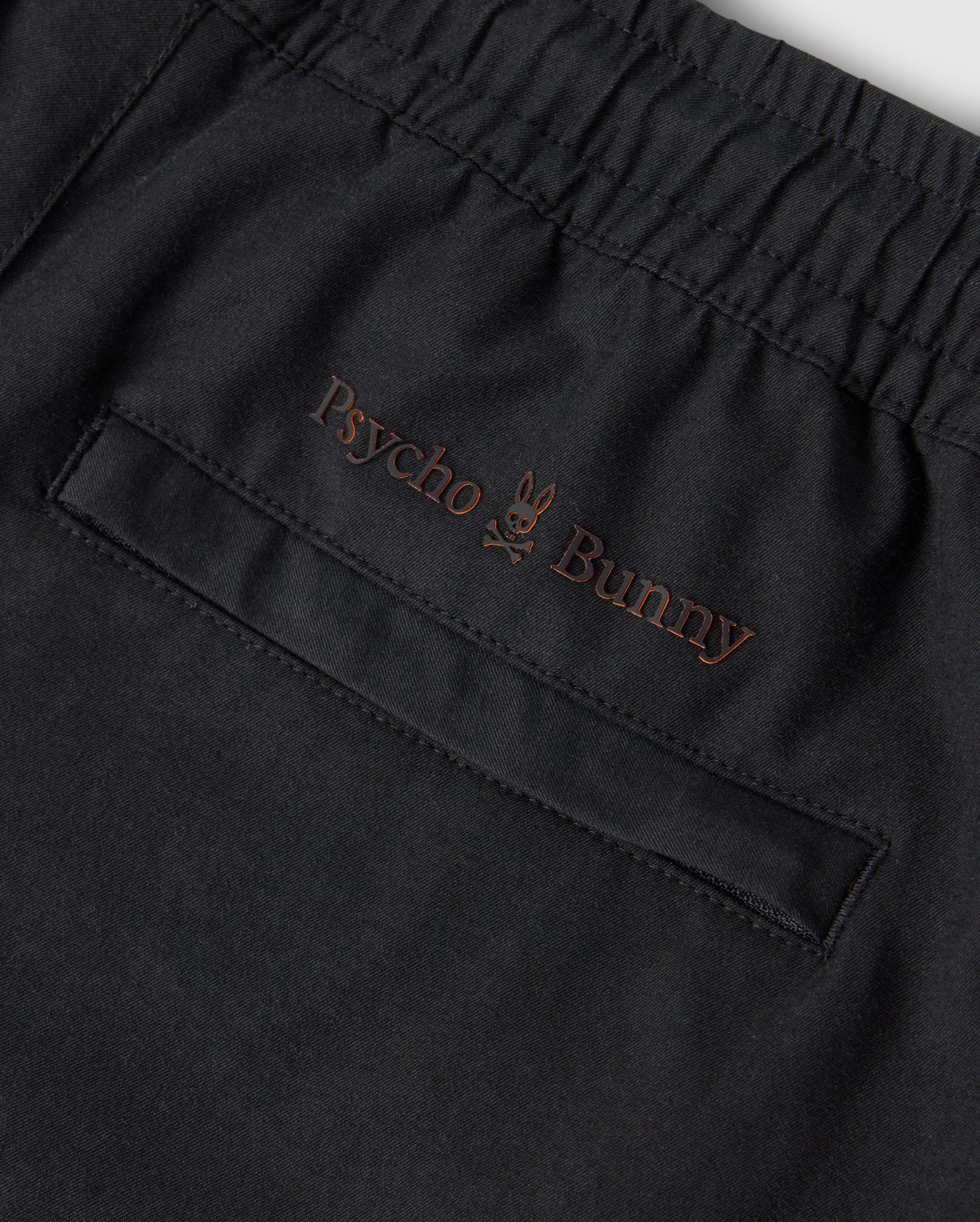 Close-up of a black KIDS MADISON COMMUTER PANT - B0P395Z1WB from Psycho Bunny, featuring an elastic waistband and a pocket with subtle brown embroidery. The embroidery includes the text "Psycho Bunny" with a small bunny logo positioned between the two words. Zippered expansion vents add functionality to this stylish piece.