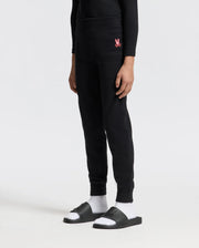 A person is standing against a plain background wearing the cozy KIDS VAL WAFFLE PANT by Psycho Bunny, in black, featuring a pink logo on the upper thigh. They are also wearing black slide sandals paired with white socks.
