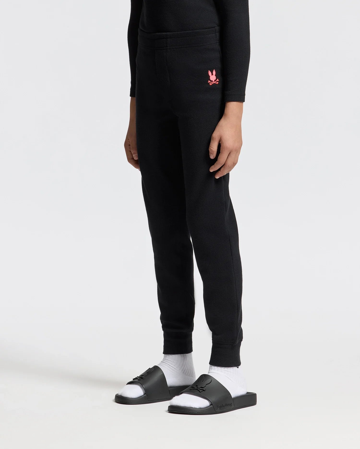 A person is standing against a plain background wearing the cozy KIDS VAL WAFFLE PANT by Psycho Bunny, in black, featuring a pink logo on the upper thigh. They are also wearing black slide sandals paired with white socks.