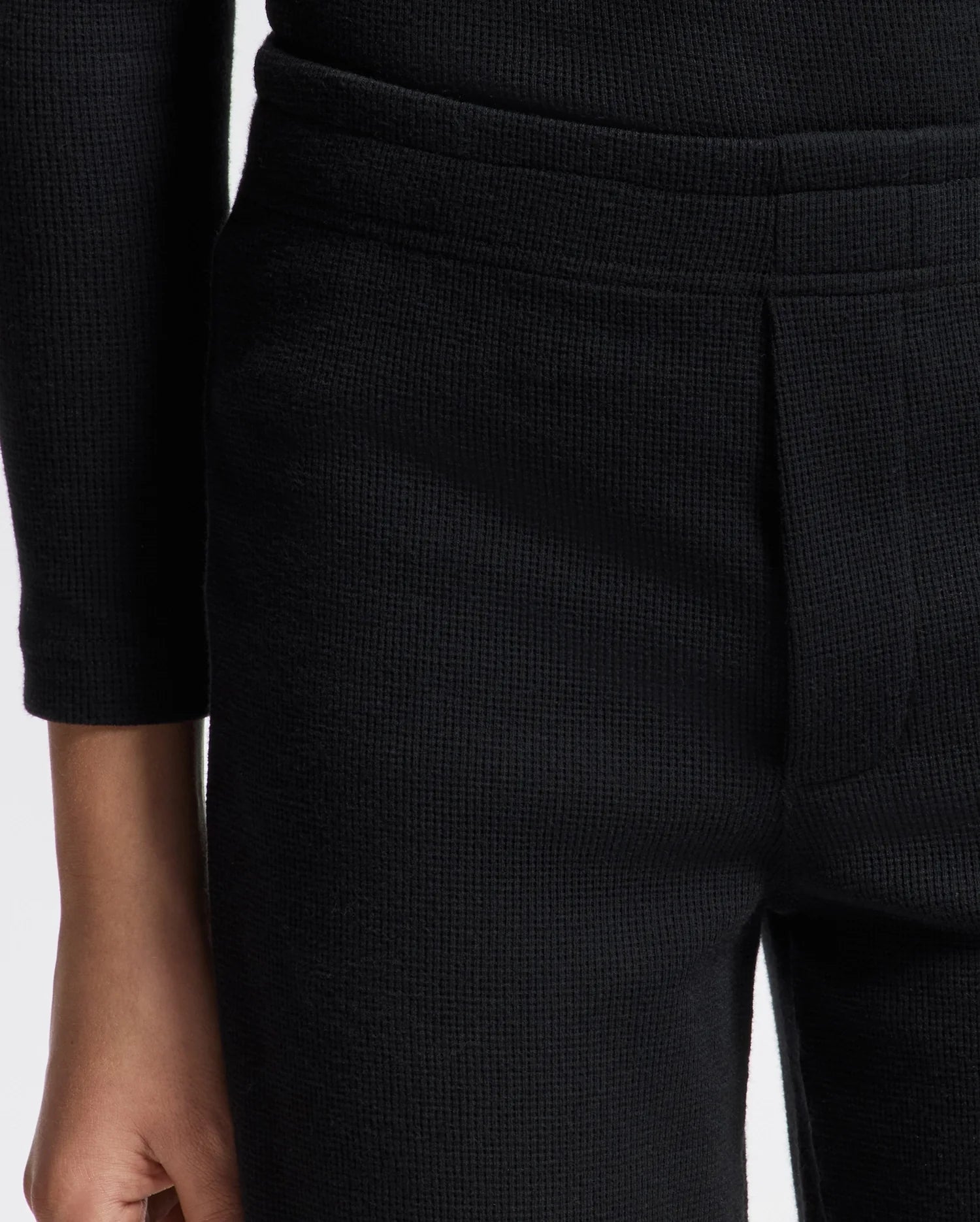A close-up shot features someone wearing a textured black garment, highlighting the waist and lower sleeve area. The material is ribbed, similar to Psycho Bunny's KIDS VAL WAFFLE PANT (B0P673E200), which offers a cozy waffle texture with a tailored fit at the waist. Only part of the person's arm and torso are visible.
