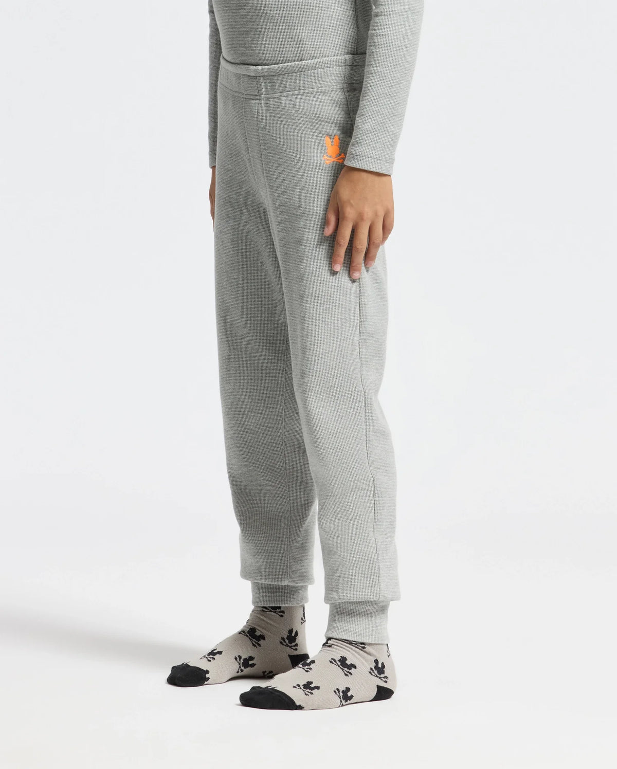 A person wearing Psycho Bunny's KIDS VAL WAFFLE PANT - B0P673E200 in light gray, matched with a coordinating long-sleeve shirt. They have one hand resting on their side. Gray socks featuring black patterns and black toe caps complete the look as they stand against a plain white background.