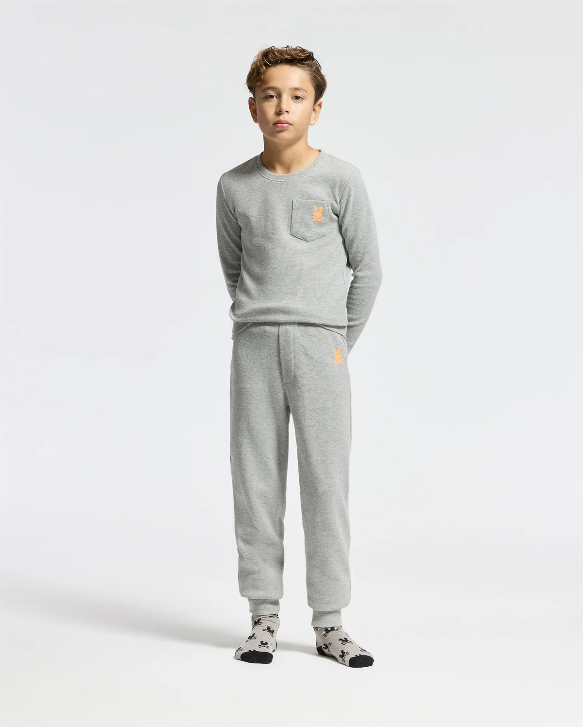 A young boy stands against a plain white background wearing the Psycho Bunny KIDS VAL WAFFLE PANT - B0P673E200, a light gray shirt and cozy waffle pants set. The outfit includes a small orange emblem on the chest and hip for a pop of color. He completes his look with patterned socks, epitomizing casual yet stylish comfort.