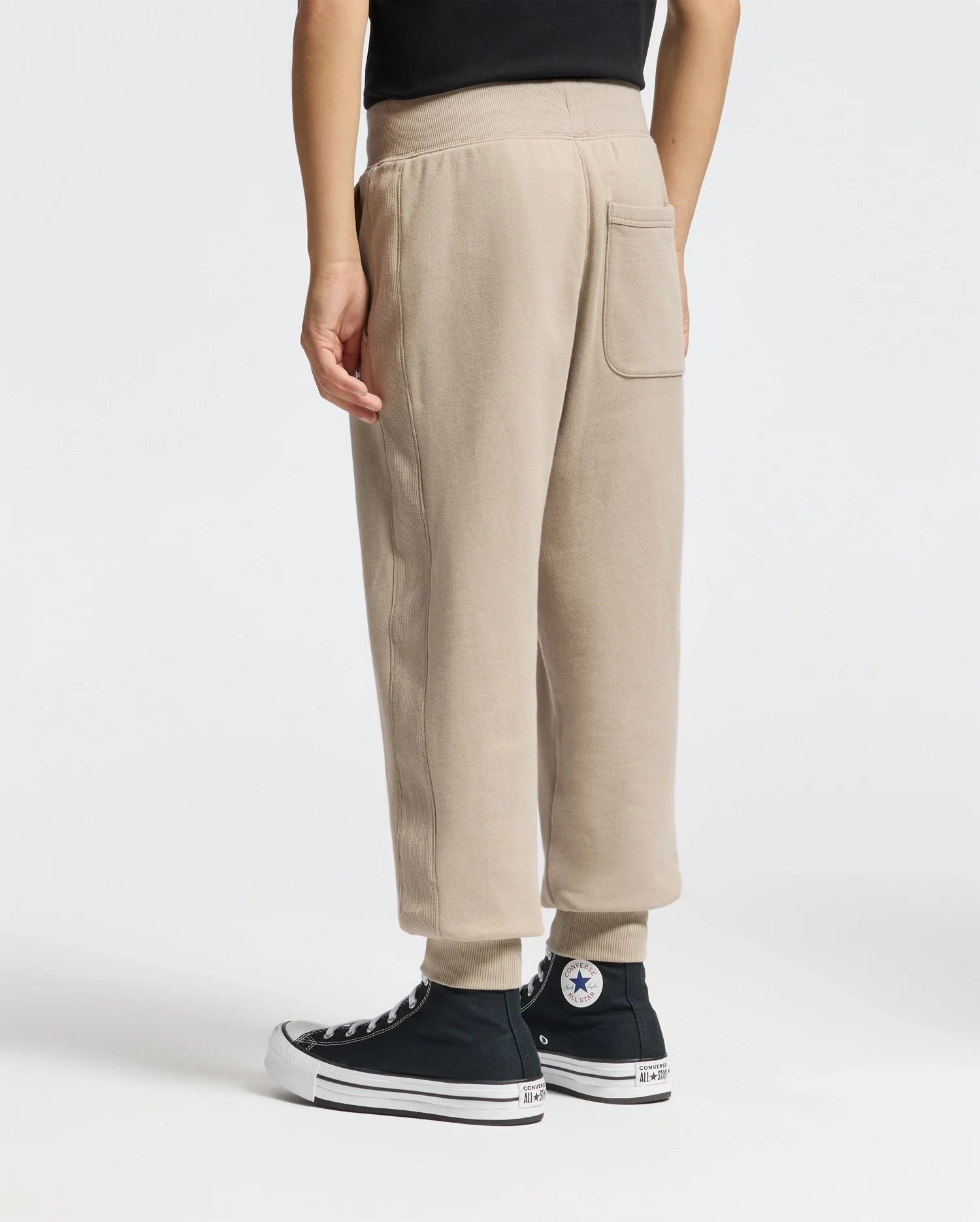 A person wearing the KIDS LEVY ESSENTIAL FLEECE SWEATPANT in beige by Psycho Bunny and black high-top Converse sneakers stands against a plain white background. Only the lower half of the individual's body is visible.