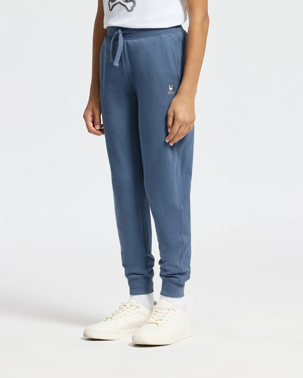 A person wearing a white T-shirt, comfortable Kids Levy Essential Fleece Sweatpants by Psycho Bunny featuring a small embroidered logo near the drawstring kangaroo pocket, white socks, and white sneakers stands against a plain background. Only the lower half of the body is visible.
