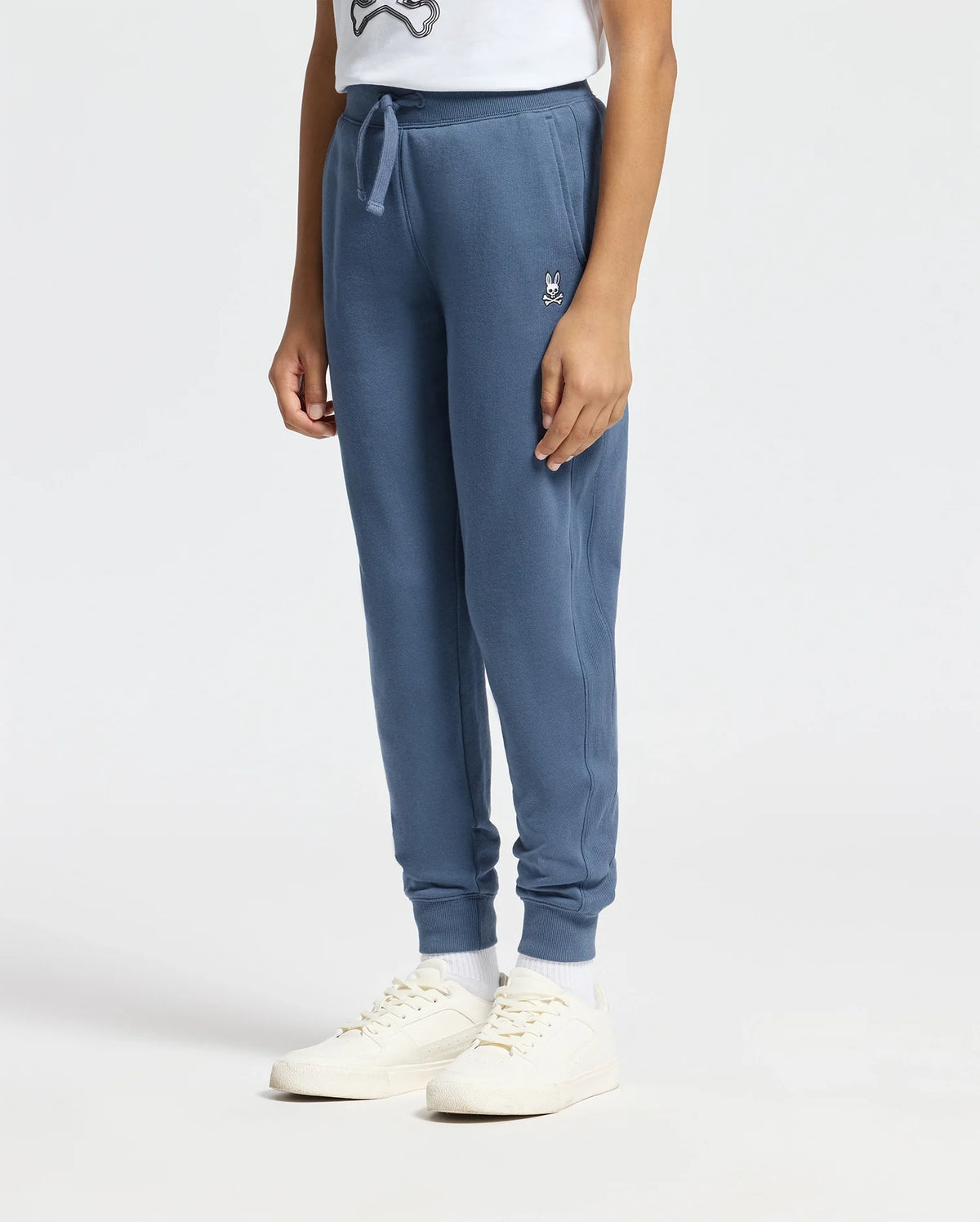 A person wearing a white T-shirt, comfortable Kids Levy Essential Fleece Sweatpants by Psycho Bunny featuring a small embroidered logo near the drawstring kangaroo pocket, white socks, and white sneakers stands against a plain background. Only the lower half of the body is visible.