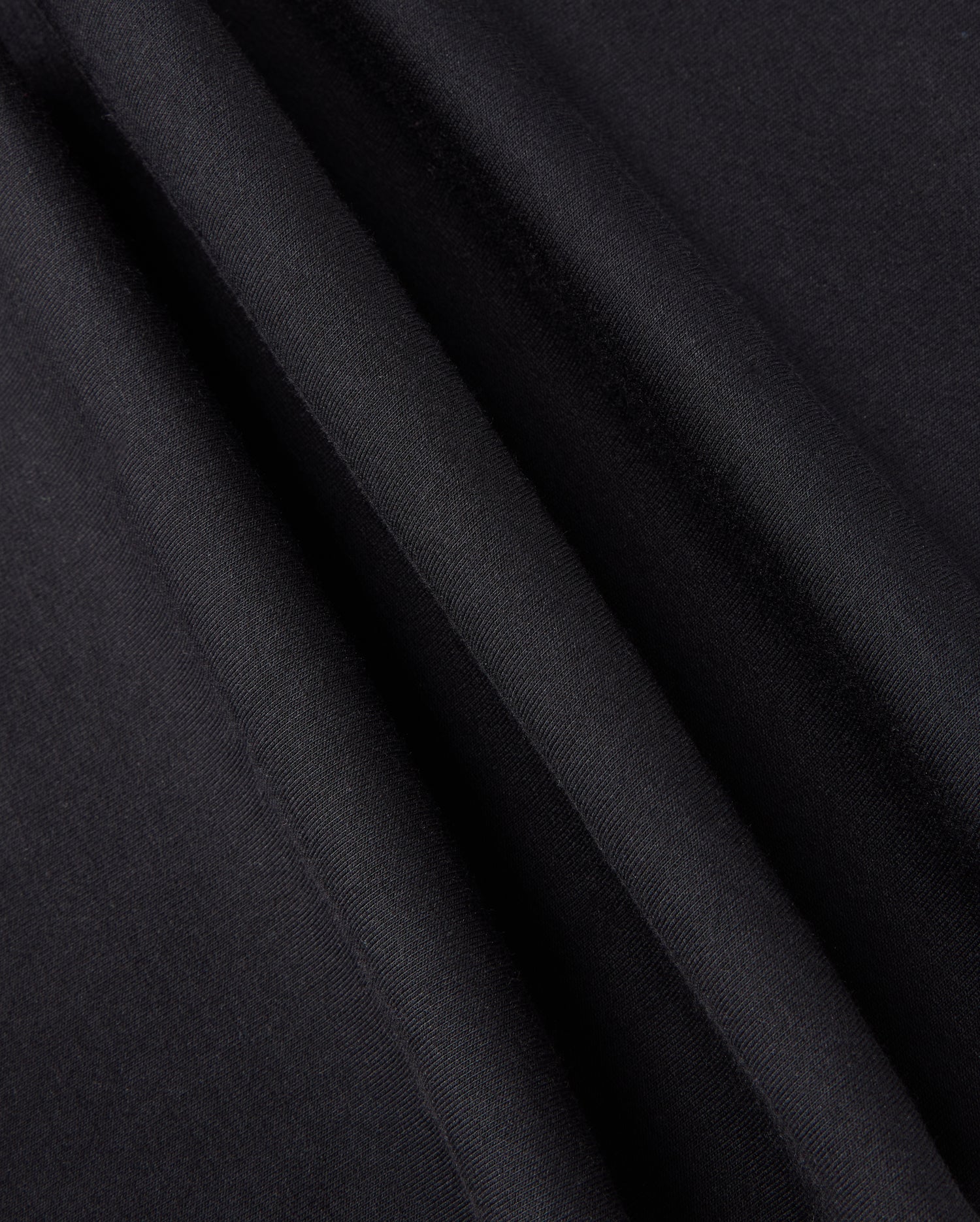 Close-up of black fabric with light reflecting off its smooth surface, creating subtle lines and textures. The material, perfect for the KIDS WALTER LIGHTWEIGHT SWEATPANT - B0P860D200 by Psycho Bunny, appears slightly folded or draped, showcasing its softness and sheen ideal for casual wear.