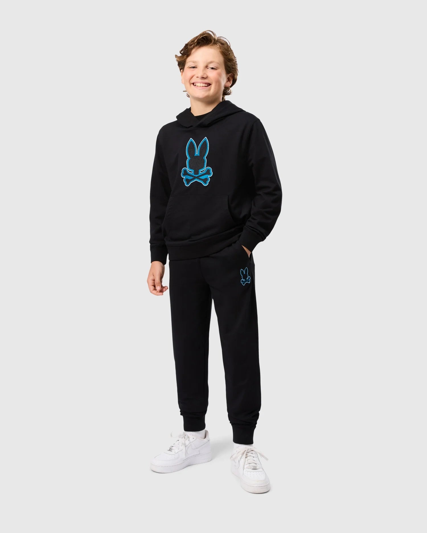 A smiling child with wavy hair stands wearing the KIDS WALTER LIGHTWEIGHT SWEATPANT - B0P860D200 black hoodie and joggers set from Psycho Bunny, featuring a blue bunny skull design. With one hand in their pocket, they showcase the casual wear outfit. The breathable fit is completed with white sneakers against a plain light gray background.