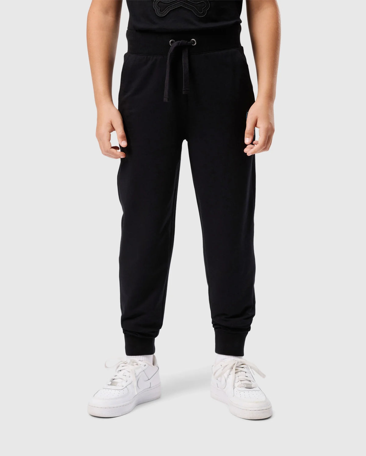 A person stands wearing the Psycho Bunny KIDS WALTER LIGHTWEIGHT SWEATPANT - B0P860D200 in black paired with white sneakers. The drawstring of the lightweight sweatpant is visible at the waist, and the person's hands are relaxed by their sides. The plain light gray background highlights the casual wear perfectly.